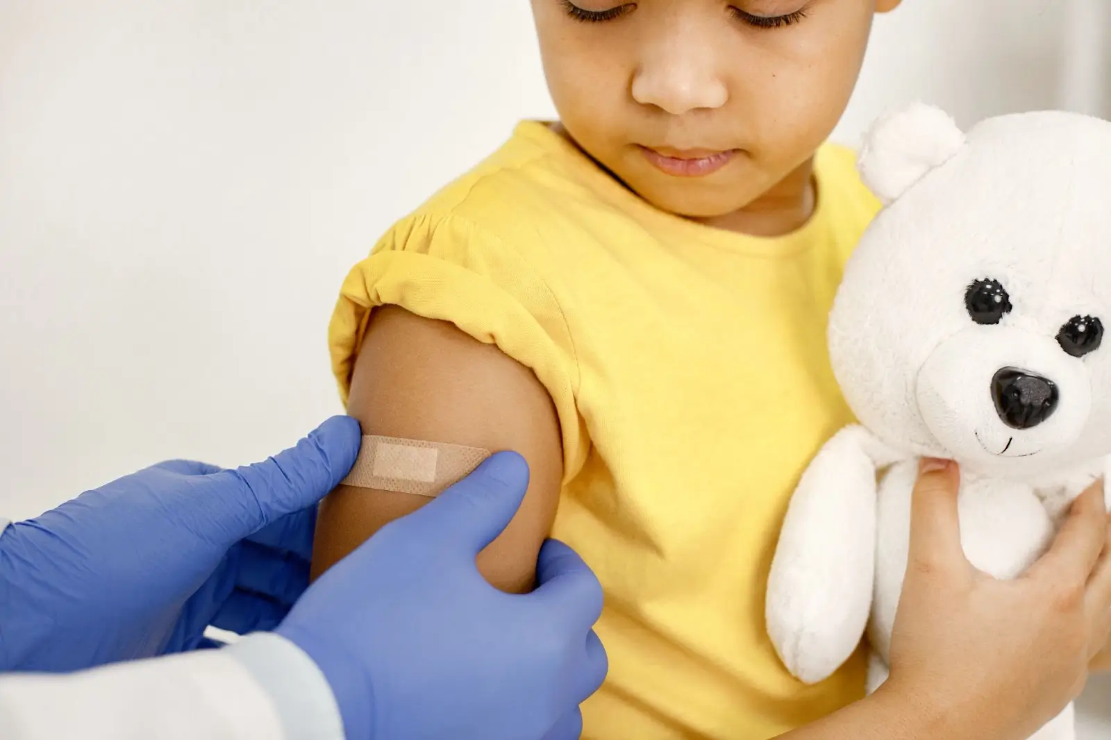A child in a yellow shirt holds a white teddy bear while a healthcare worker with blue gloves applies a bandage to the child’s upper arm after vaccination.