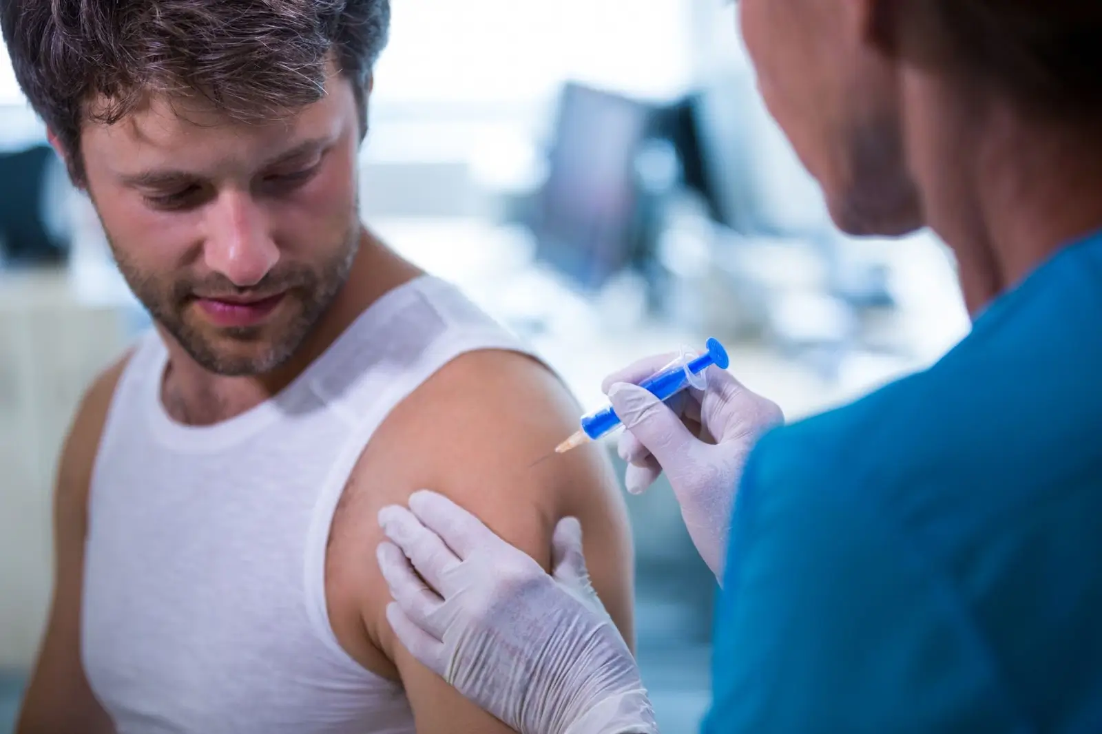 A man in a white tank top receives a vaccine injection in his upper arm from a healthcare worker wearing gloves and blue scrubs in a clinical setting.