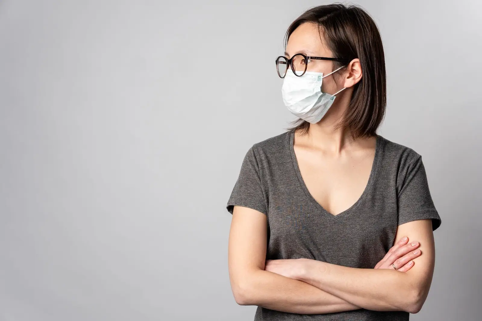 A woman wearing glasses and a medical face mask stands with her arms crossed, looking to the side. She has shoulder-length brown hair and is wearing a grey t-shirt, with a neutral grey background behind her.
