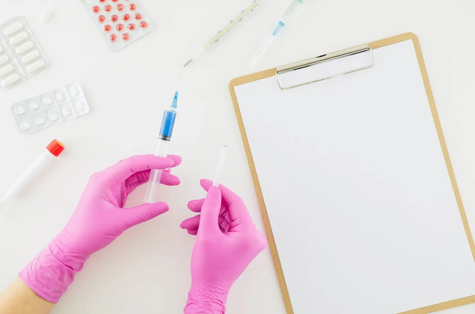 A person wearing pink gloves prepares a syringe with medicine beside a blank clipboard, pills, and other medical supplies on a white surface.