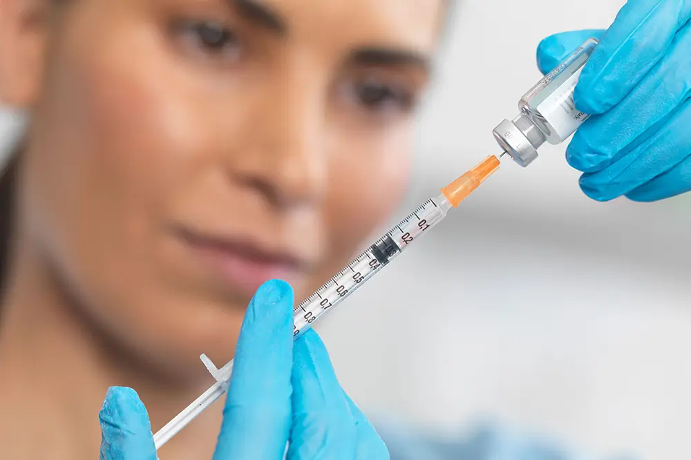 A person wearing blue gloves draws liquid from a vial into a syringe, with their face softly blurred in the background.