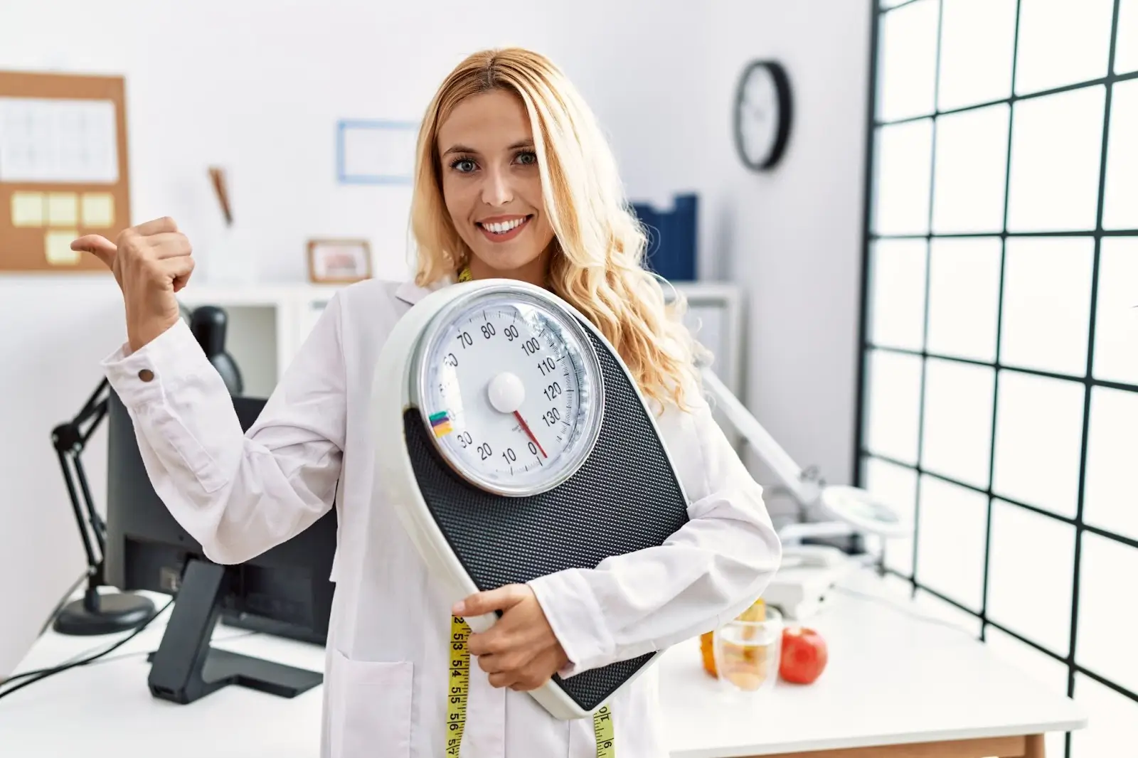 A smiling woman in a white coat holds a bathroom scale and gestures with her thumb. In the background, there's a desk with fruits, measuring tape, and a bright window, suggesting a health or nutrition setting.