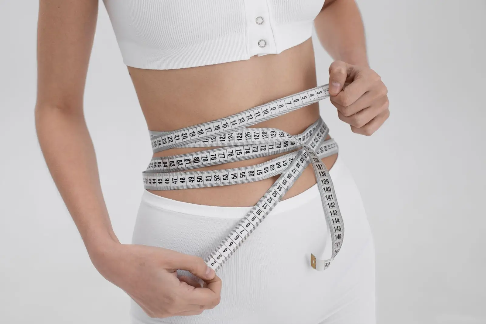 A person in white clothes measures their waist with a measuring tape, highlighting a focus on fitness or weight management. The background is plain and light-colored.