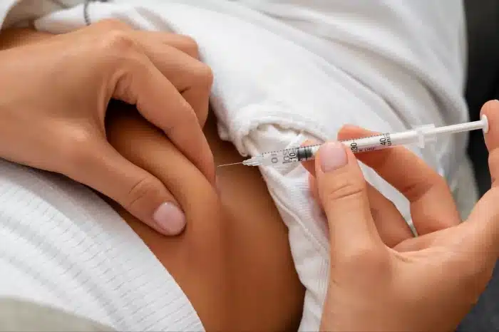 A person gets an injection in the abdomen with a syringe, with the clinician pulling up their shirt and pinching the skin to administer the shot.