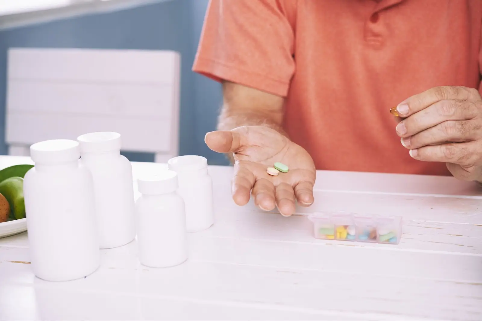 A person in an orange shirt holds several pills in one hand over a table with pill bottles and a weekly pill organizer.