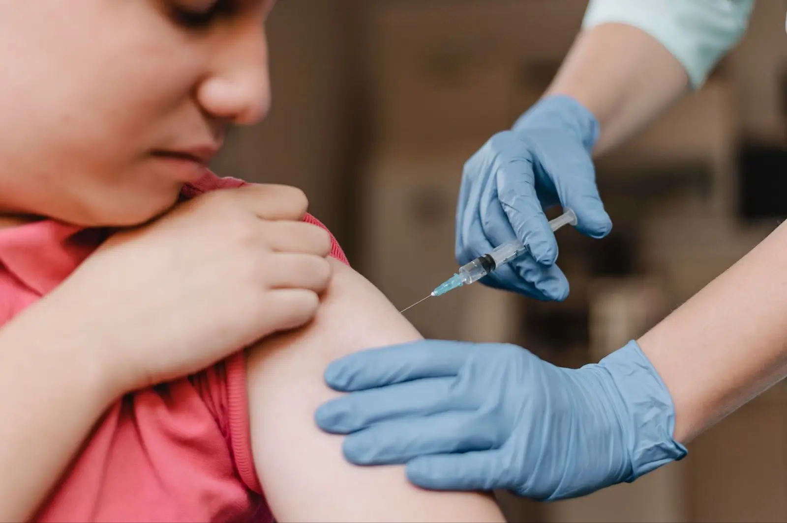 A person in a pink shirt receives a vaccine injection in their upper arm from a healthcare worker wearing blue gloves. The focus is on the injection and gloved hands.