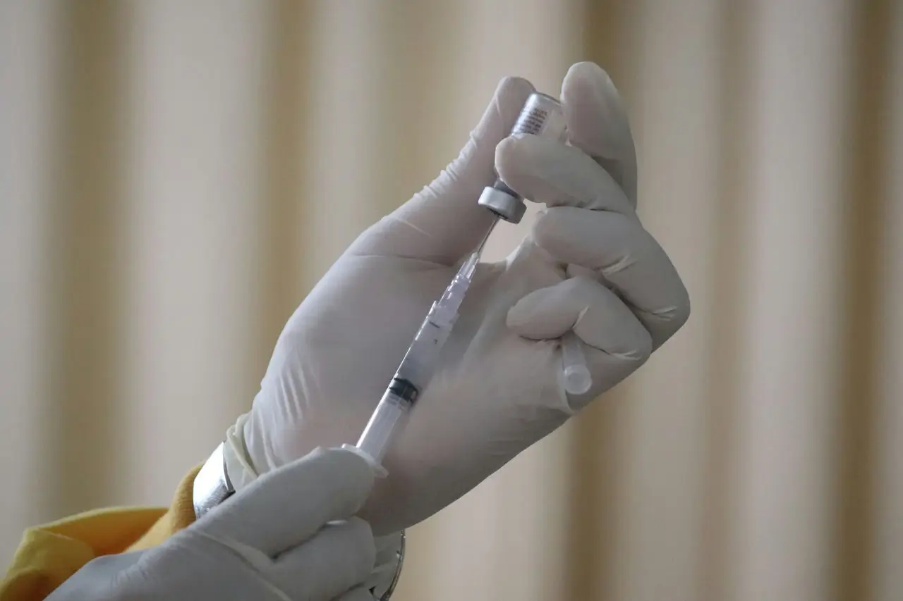 A gloved hand holds a vial and a syringe, drawing liquid from the vial against a blurred beige background, suggesting a medical or vaccination setting.