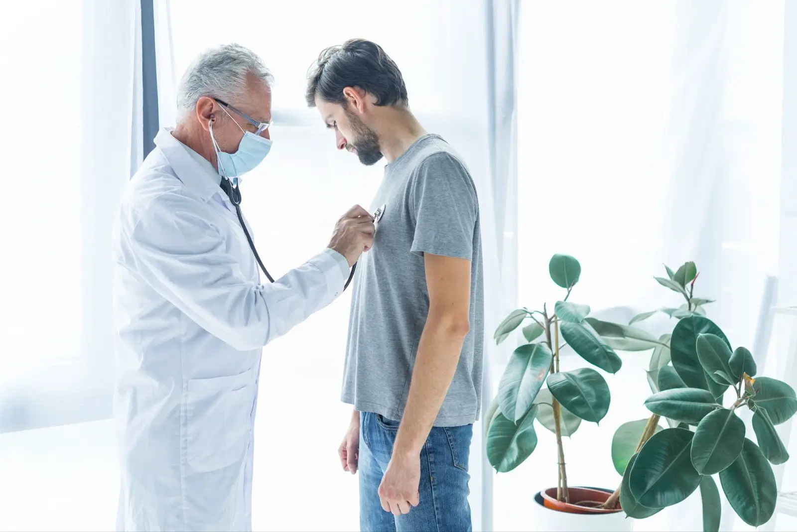A doctor wearing a white coat and face mask uses a stethoscope to listen to the chest of a man in a gray t-shirt during a medical examination in a bright room with potted plants.