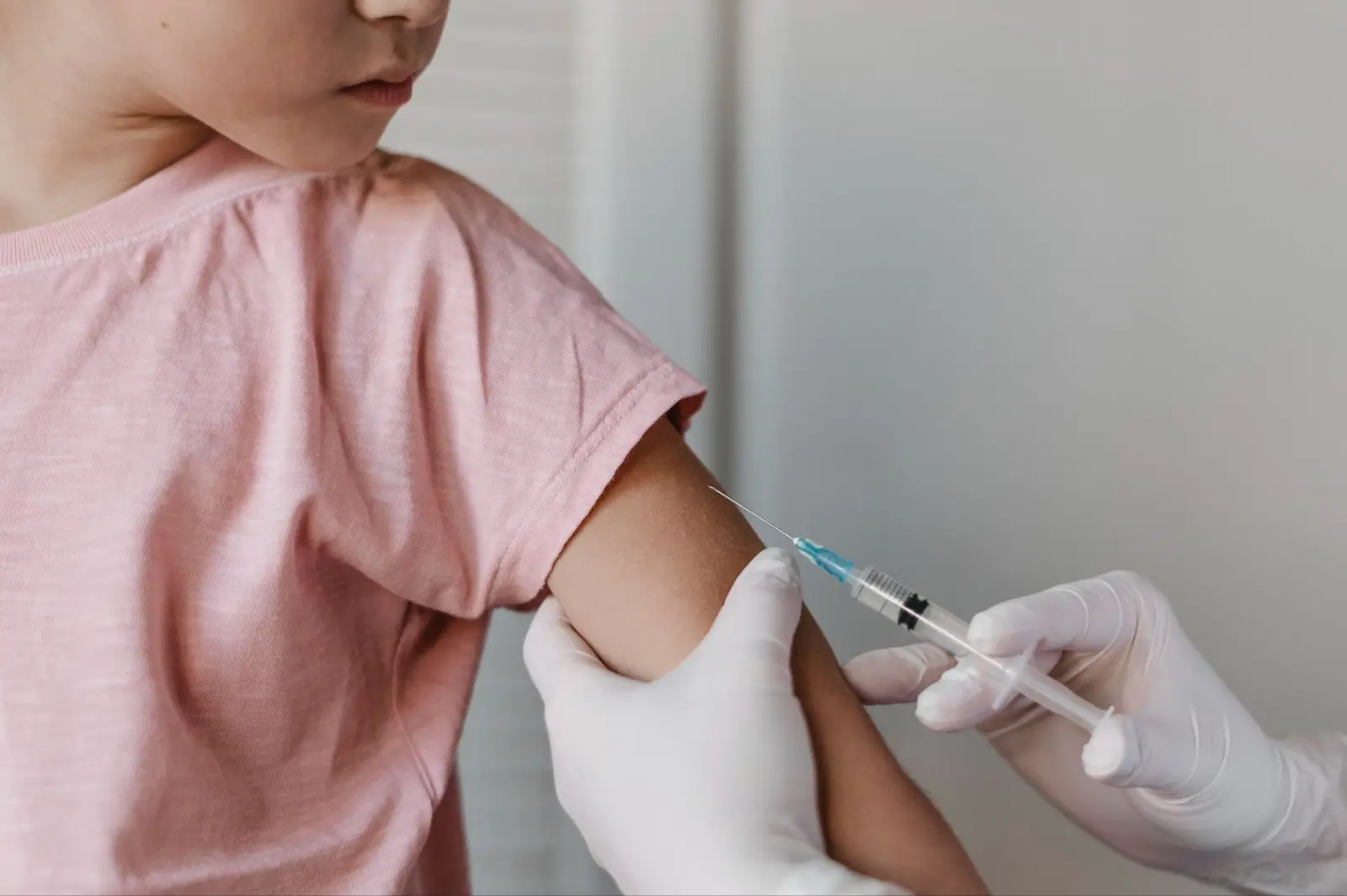 A healthcare worker wearing white gloves administers a vaccine injection to a child's upper arm. The child is wearing a pink shirt and looking away from the needle.
