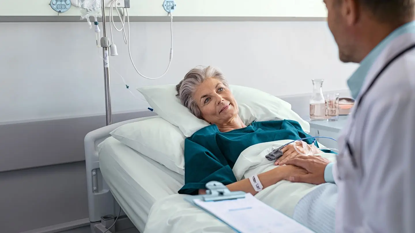An older woman lies in a hospital bed, wearing a gown, looking at a doctor who is sitting beside her and holding her hand. Medical equipment and an IV drip are visible in the background.