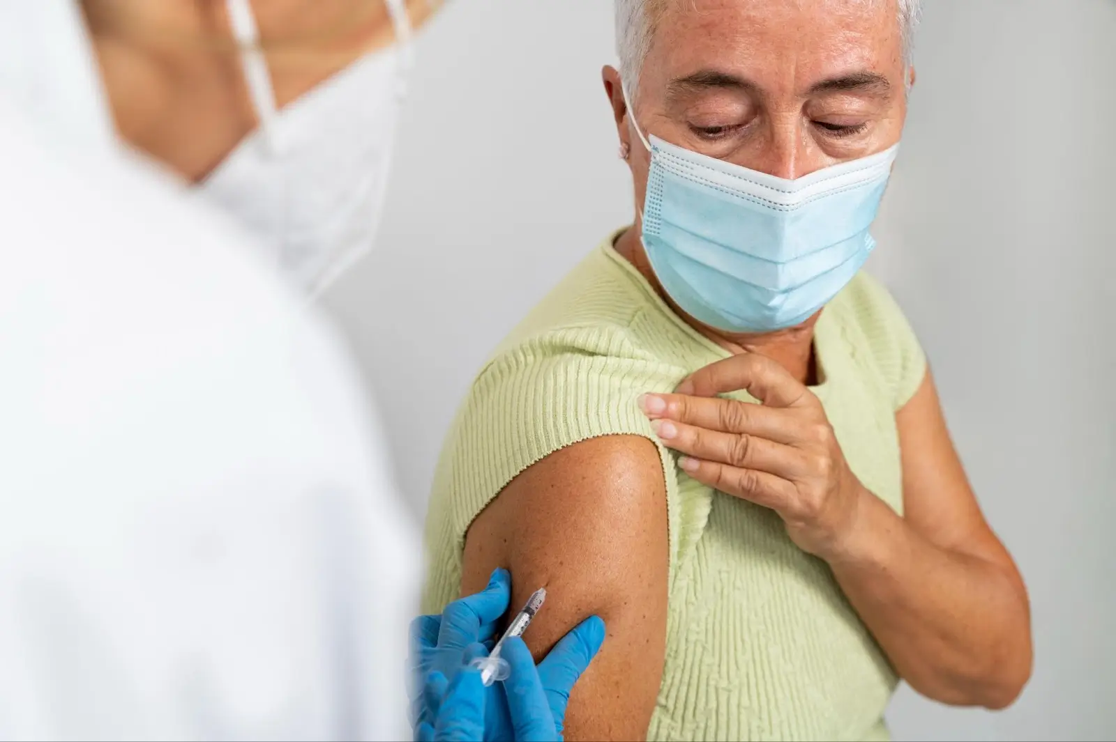 An older adult wearing a mask pulls up their sleeve as a healthcare worker, also masked and gloved, prepares to administer a vaccine injection in their upper arm.