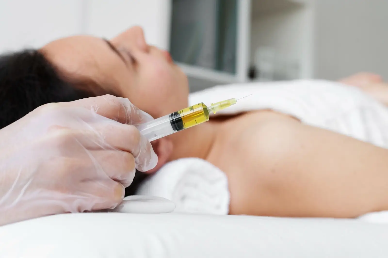 A person lying down in a white robe, with a gloved hand holding a syringe filled with yellow liquid near their shoulder, preparing for an injection in a clinical setting.
