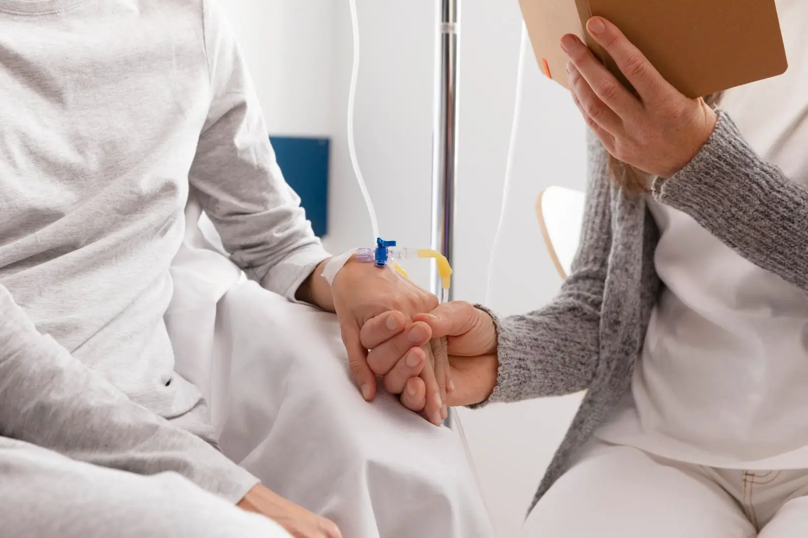 A person in a hospital bed with an IV in their hand is being comforted by another person who is holding their hand and a notebook. Both individuals are wearing light-colored clothing.