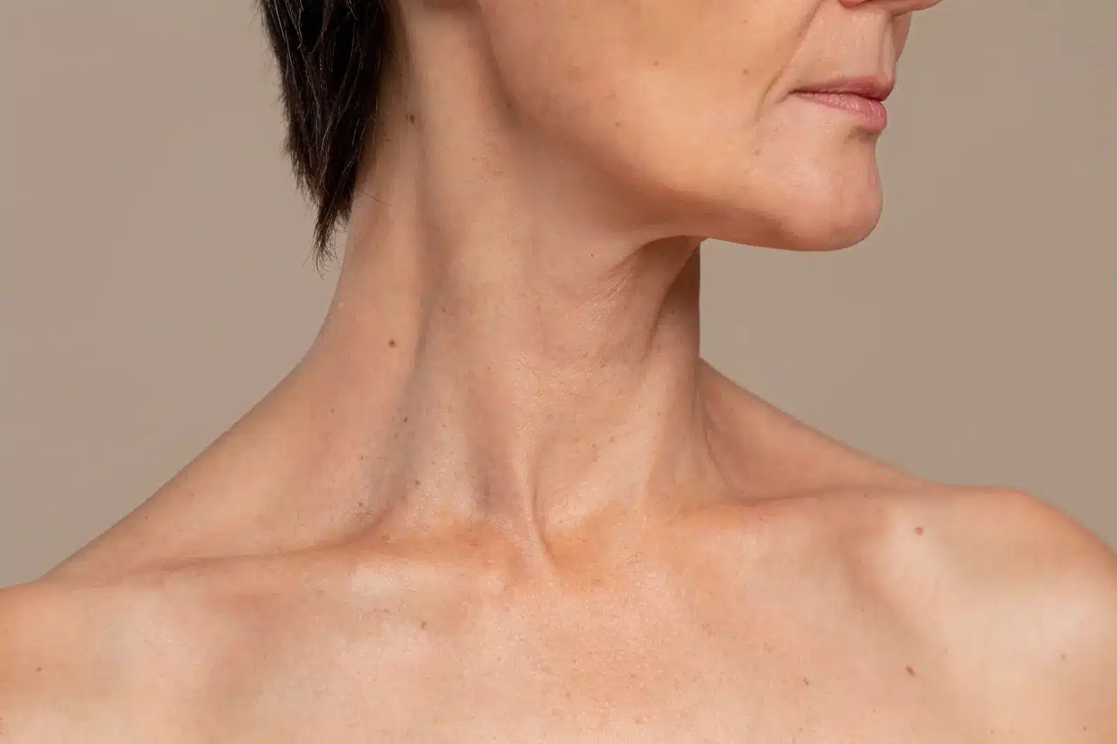 Close-up of a middle-aged person's neck, jawline, and upper chest with short dark hair, visible skin texture, and subtle signs of aging. The background is a plain, neutral color.