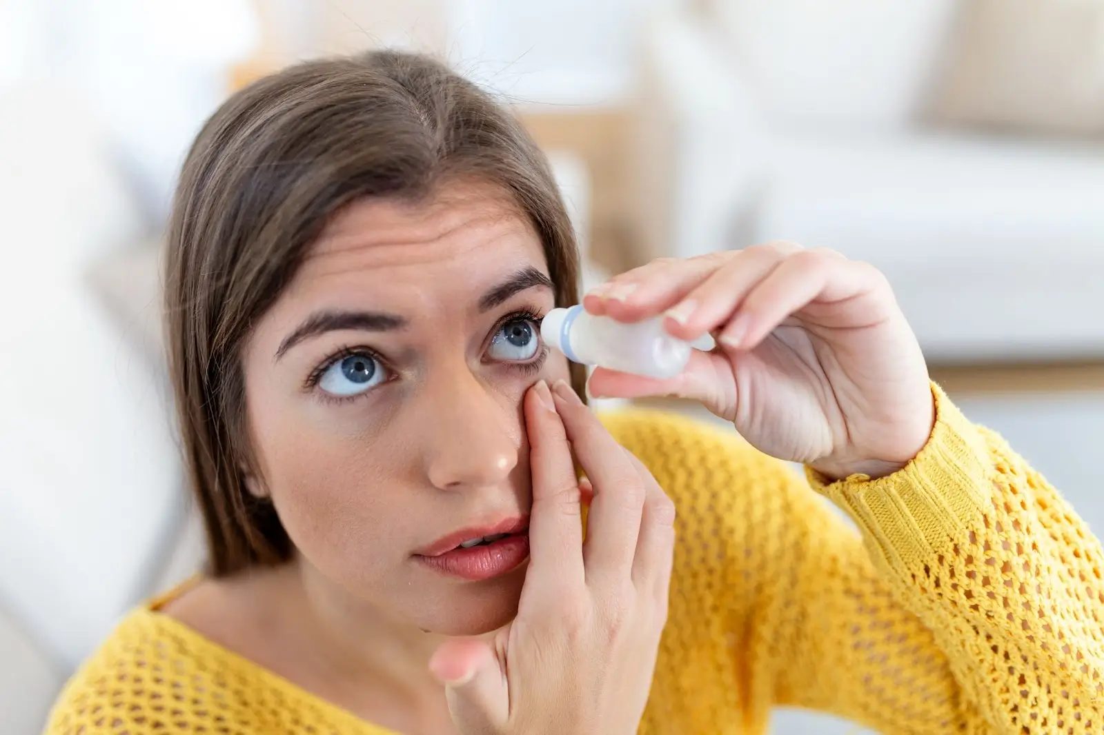 A woman in a yellow sweater tilts her head back and applies eye drops to her right eye while holding her lower eyelid down.