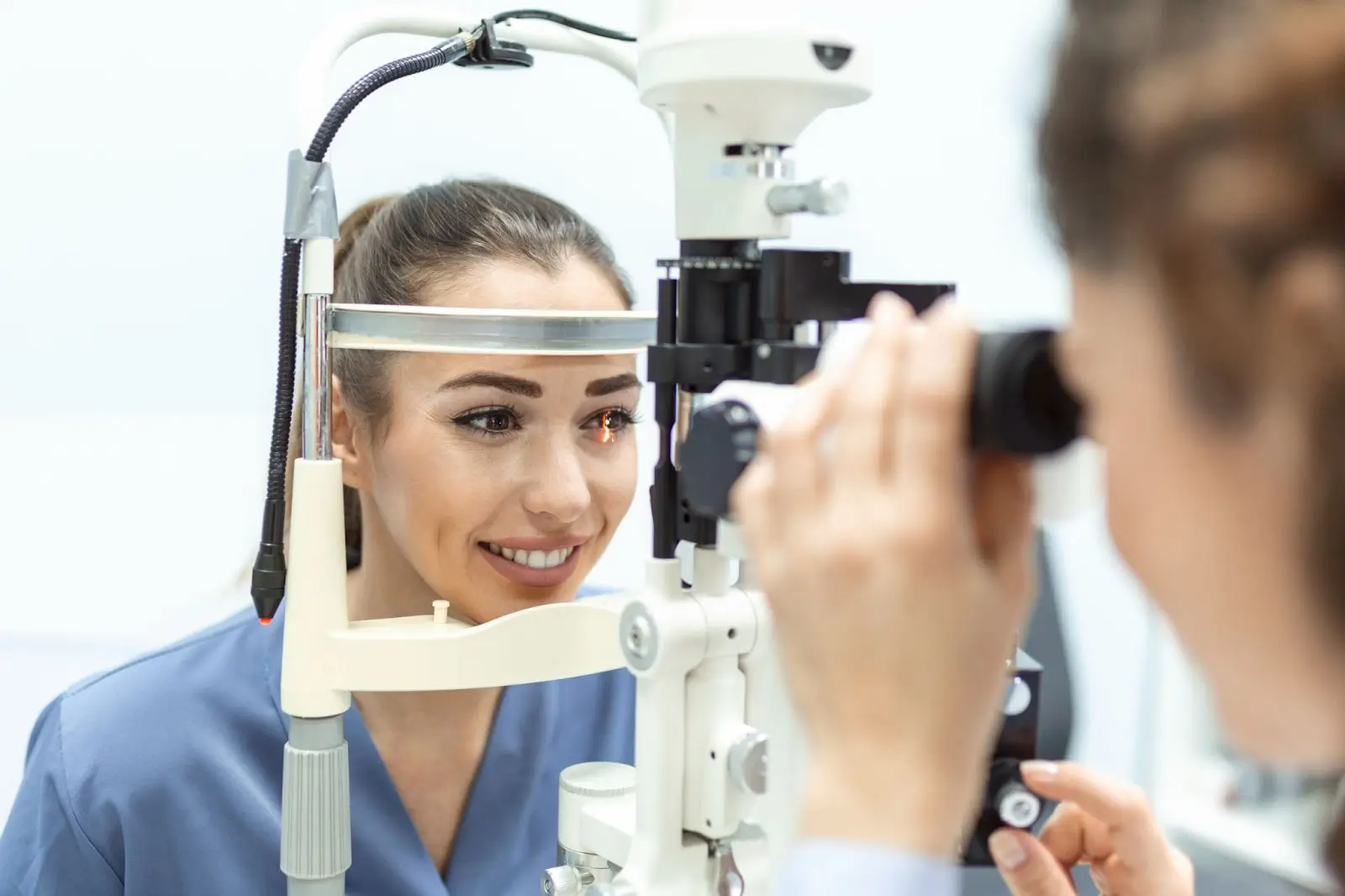 A woman is having her eyes examined with a slit lamp by an eye care professional in a clinic. The patient is smiling, and the examiner is adjusting the instrument to check her eyes.