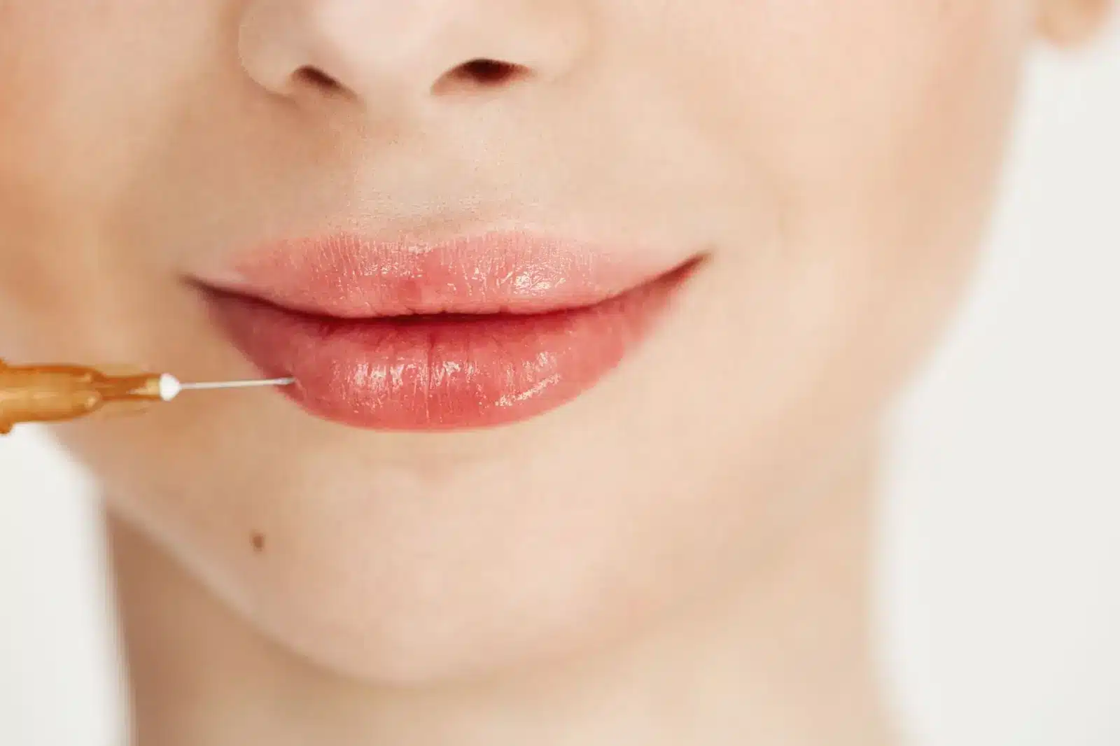A close-up of a woman's lower face with smooth skin and glossy lips. A syringe is positioned near her lower lip, suggesting a cosmetic procedure such as a lip filler injection.