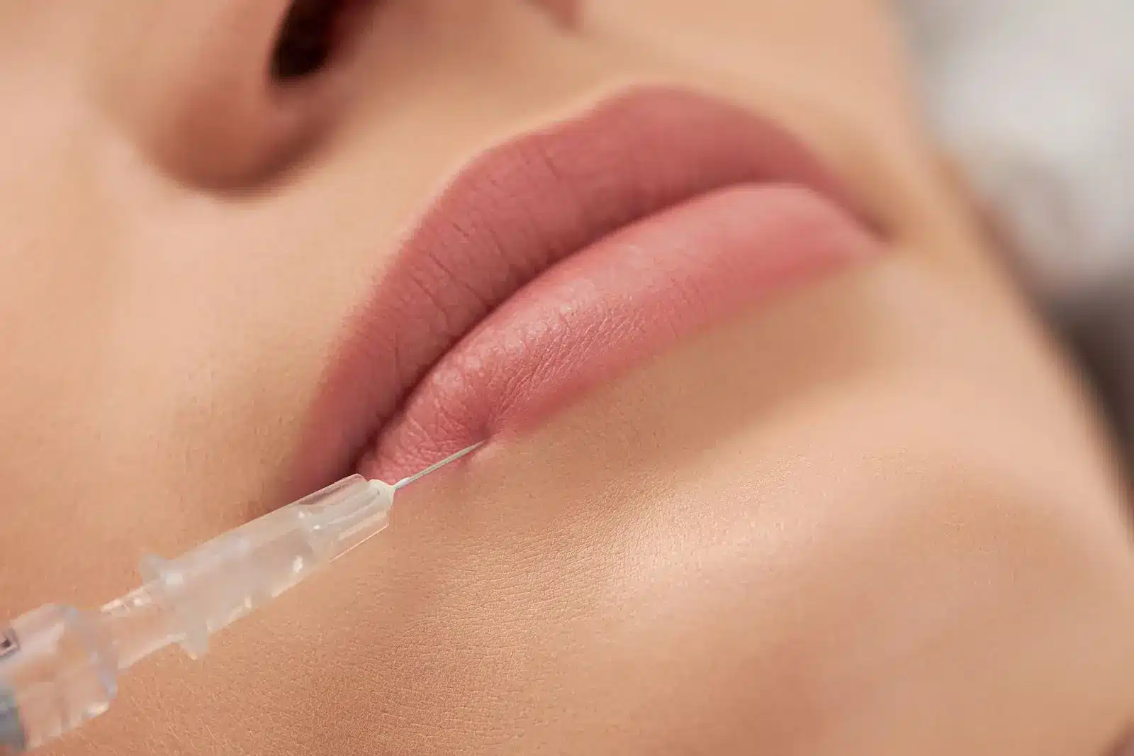Close-up of a person’s lips as a syringe, likely for a cosmetic injection or lip filler, is gently inserted into the lower lip area. The skin and lips appear smooth and healthy.