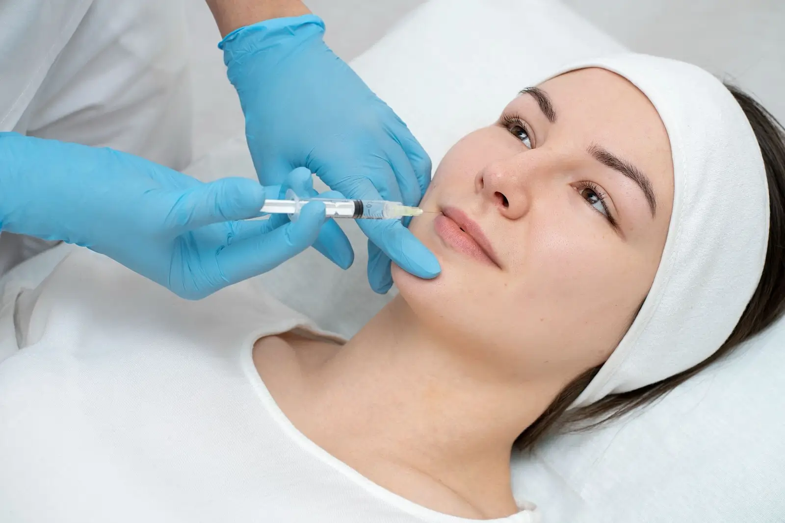 A woman lying on a treatment bed receives a cosmetic injection in her face from a medical professional wearing blue gloves. The woman wears a white headband and shirt, and appears calm during the procedure.