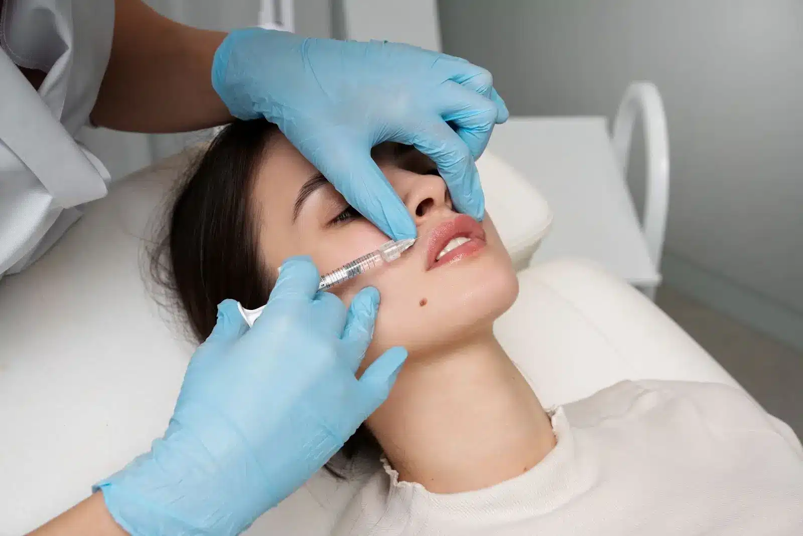 A person wearing blue gloves injects a needle into a woman's lips as she lies on a medical chair, possibly receiving a cosmetic procedure such as lip fillers or an injectable treatment.