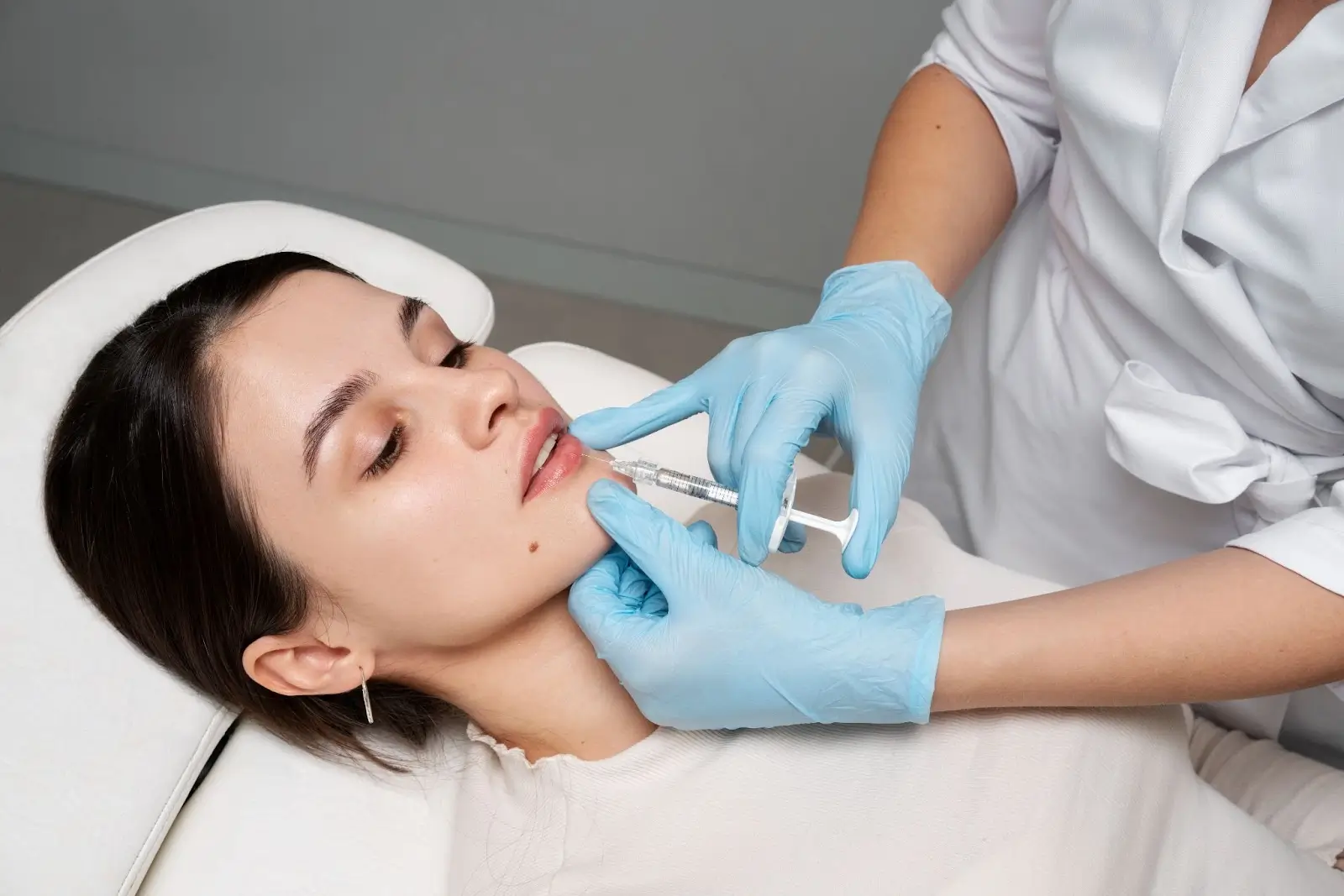 A woman lying on a clinic bed receives a lip injection from a medical professional wearing blue gloves and a white coat. The procedure appears to be cosmetic, possibly for lip enhancement or filler.