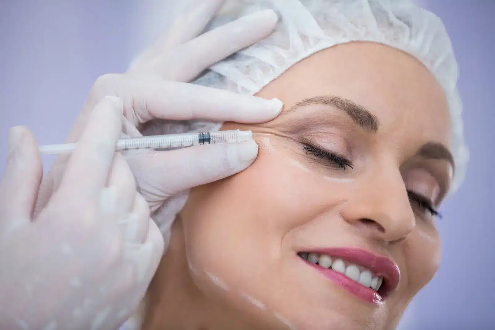 A woman wearing a hair cap smiles with her eyes closed while a gloved medical professional injects a syringe near her eye, possibly administering a cosmetic treatment like Botox.