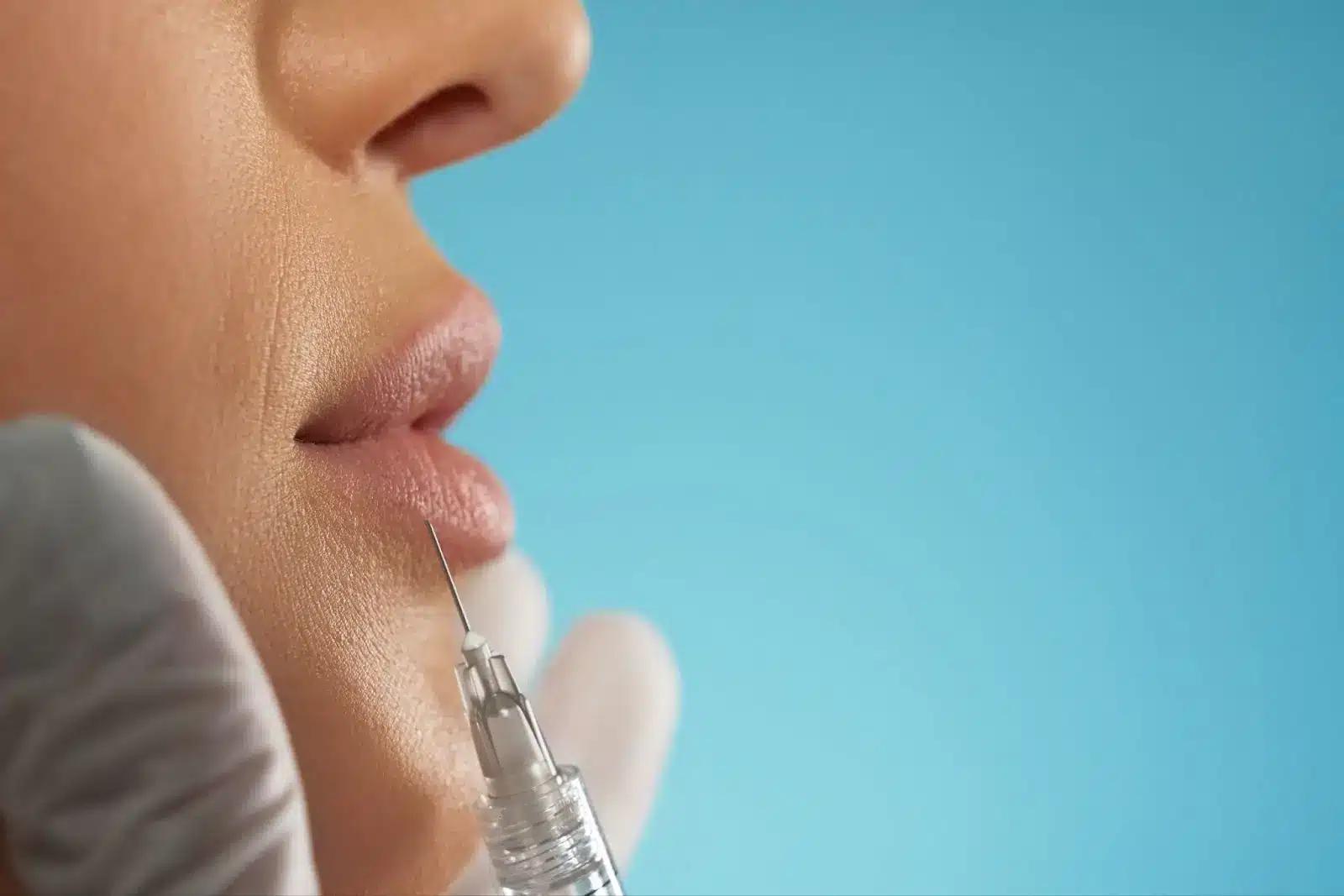A close-up of a person's lips receiving an injection, likely for a cosmetic procedure, with a gloved hand holding the syringe against a blue background.