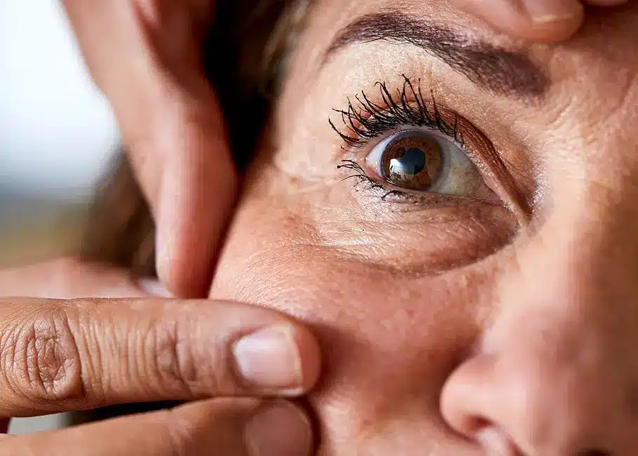A close-up of a person’s face, focusing on one eye. Fingers gently pull down the lower eyelid, revealing the eye and surrounding skin with fine lines and visible eyelashes.