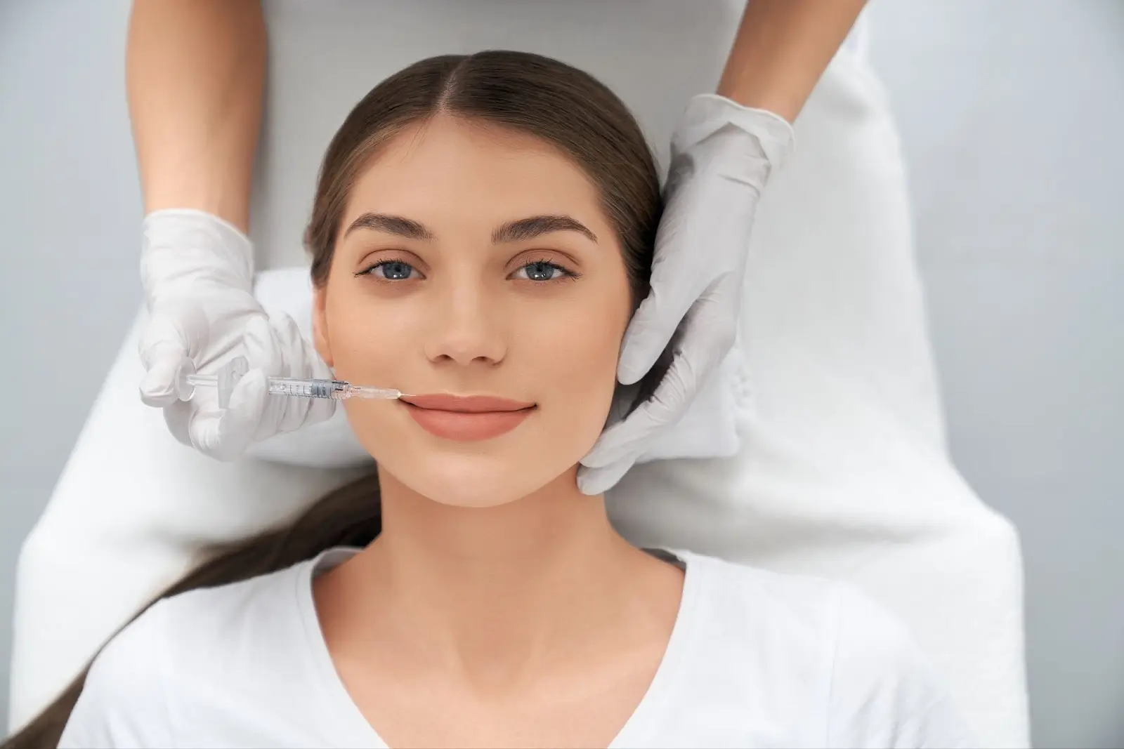 A woman lies on a medical chair while a gloved practitioner prepares to give her a facial injection with a syringe near her lips. She has long brown hair and is wearing a white top.