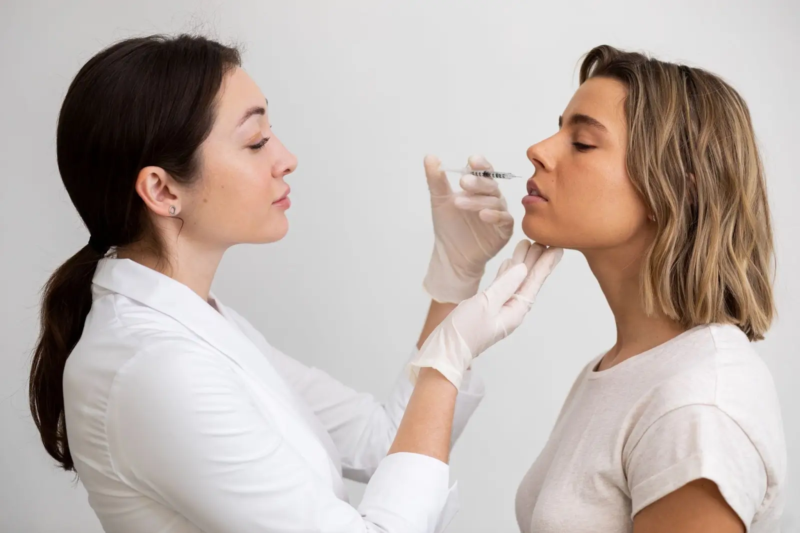 A female doctor in a white coat and gloves prepares to inject a syringe into the lips of a young woman with short hair, who is wearing a white t-shirt and tilting her face slightly upward.