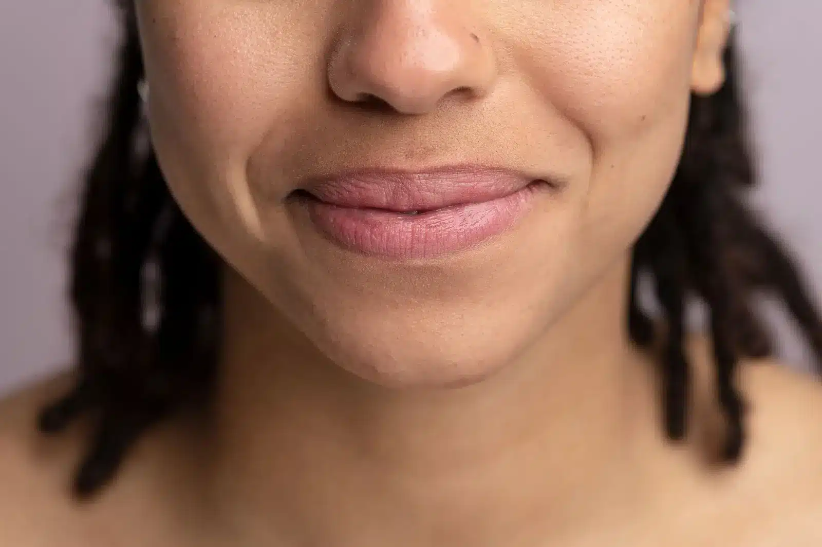 Close-up of a person's mouth and lower face, showing a subtle smile and dimples, with medium brown skin and a small nose ring. The background is blurred, and some dark hair is visible at the sides.