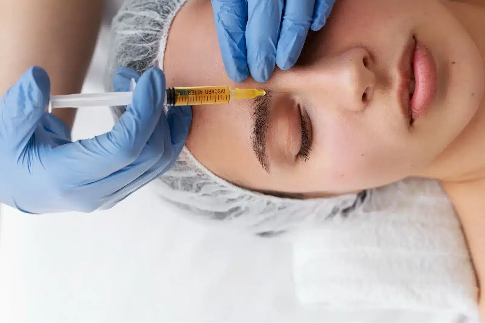 A person with closed eyes lies on a towel while someone wearing blue gloves prepares to inject a syringe into the person's forehead. The person wears a disposable cap, suggesting a clinical or cosmetic procedure.