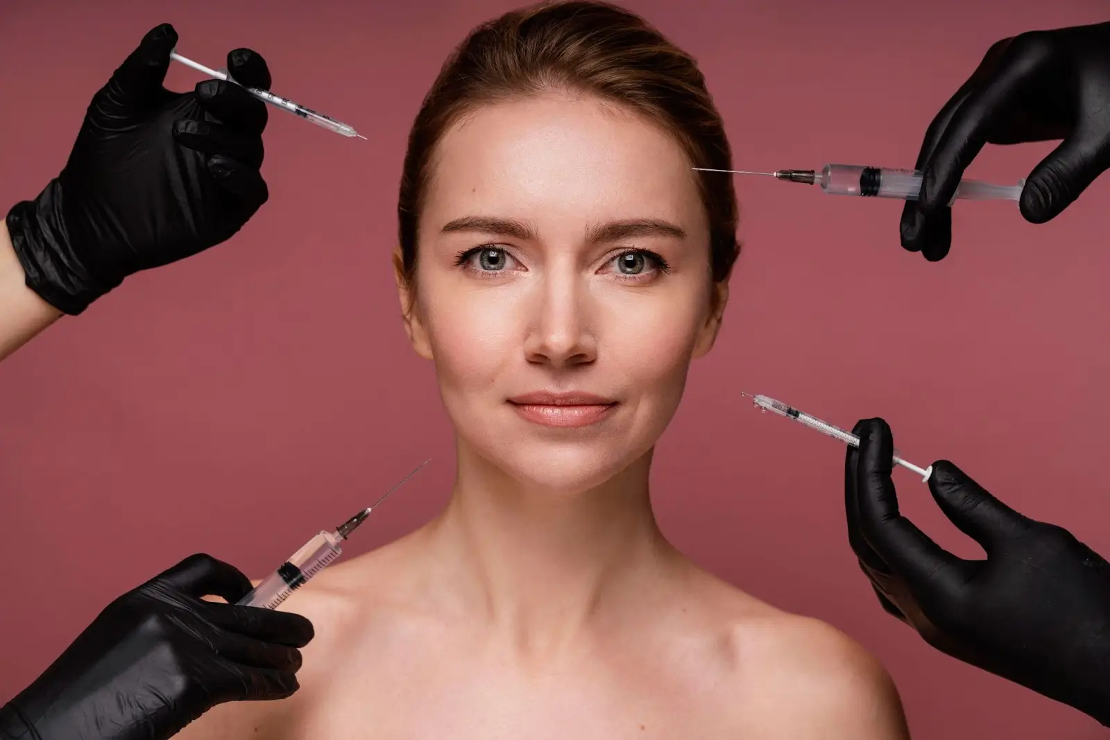 A woman with bare shoulders faces the camera against a pink background, while three gloved hands holding syringes are positioned near her face, suggesting cosmetic procedures.