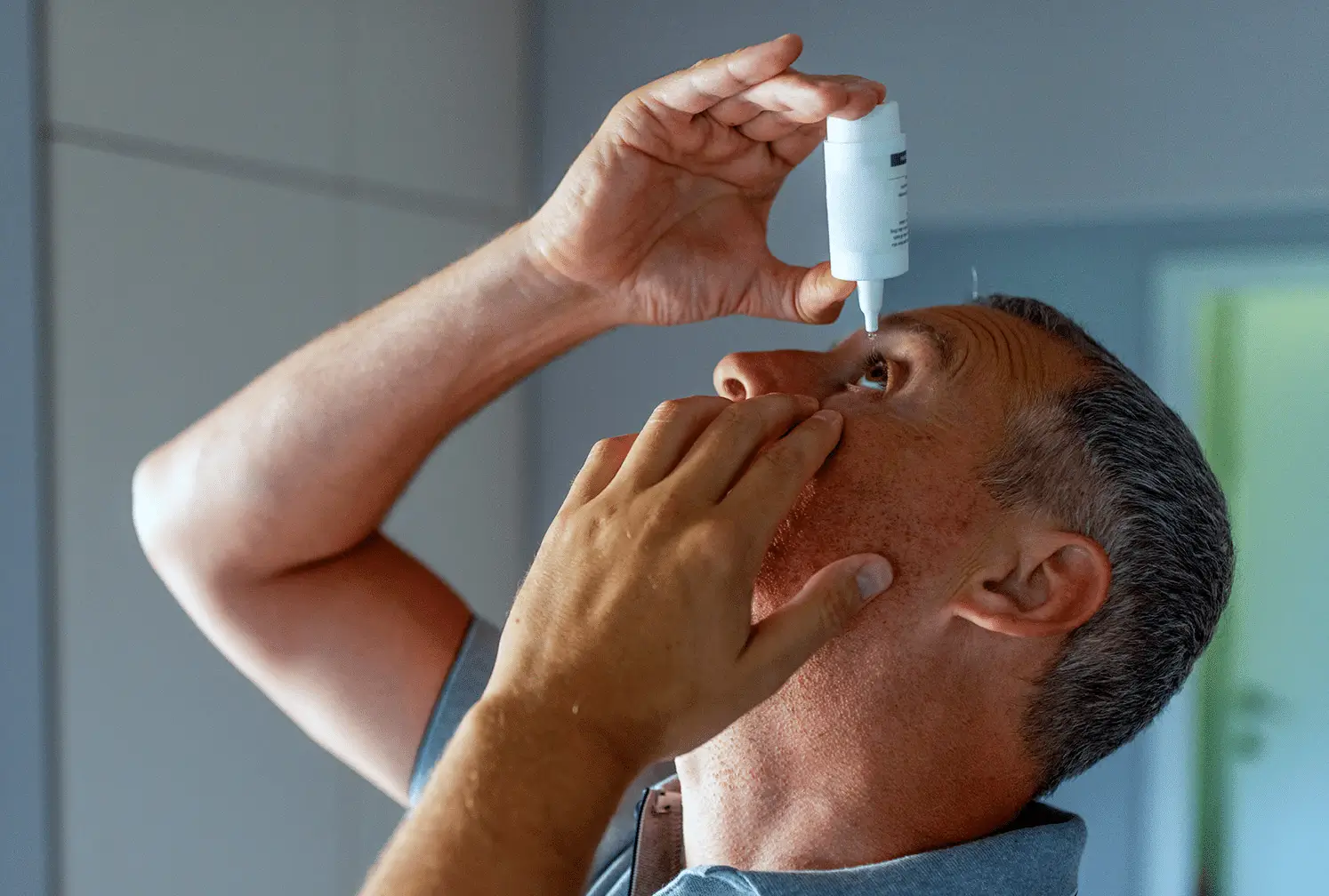 A man tilts his head back and uses one hand to hold open his eye while squeezing eye drops from a bottle into his eye.