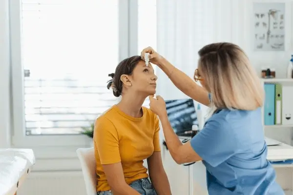 A healthcare professional in blue scrubs administers eye drops to a woman in a yellow shirt, who is seated and looking up, inside a bright medical office.