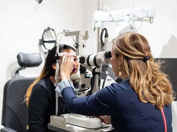 An optometrist examines a patient’s eyes using a slit lamp microscope in a bright, modern eye clinic. The patient is seated with her chin on the device while the optometrist looks through the eyepiece.