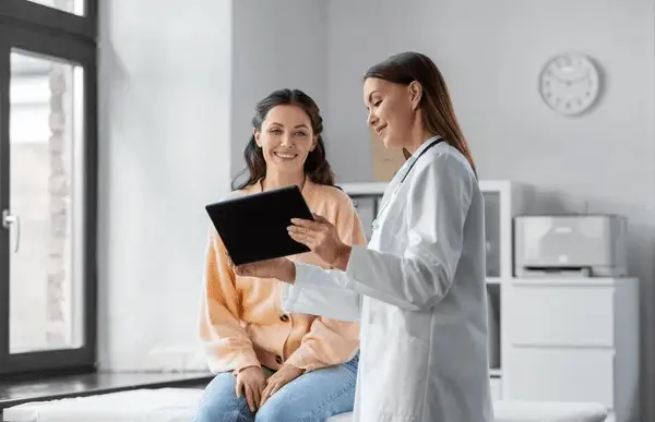 A doctor in a white coat shows a digital tablet to a smiling woman sitting on an exam table in a bright medical office.