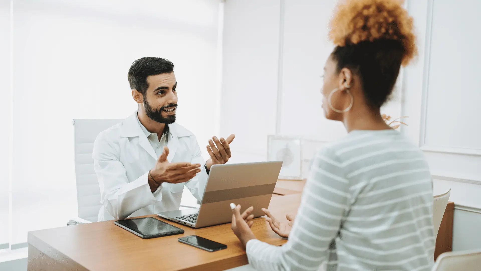 A doctor in a white coat sits at a desk, talking to a patient. Both are engaged in conversation. The doctor gestures with his hands, and there is a laptop, tablet, and phone on the desk.