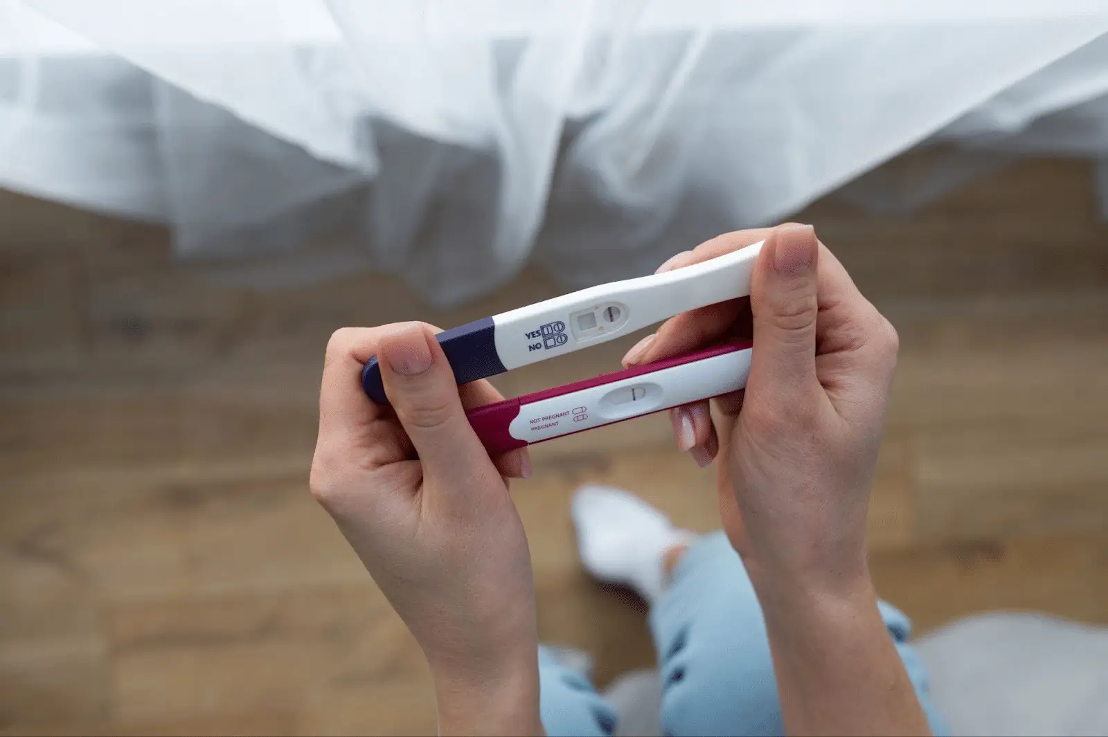 A person holding two pregnancy tests, both showing negative results, with a blurred background of light curtains and a wooden floor.