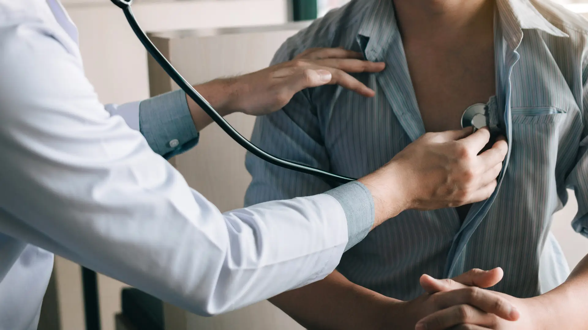 A doctor uses a stethoscope to listen to a patient's chest, who has opened their shirt for the examination. The doctor's hand holds the stethoscope against the patient's skin.