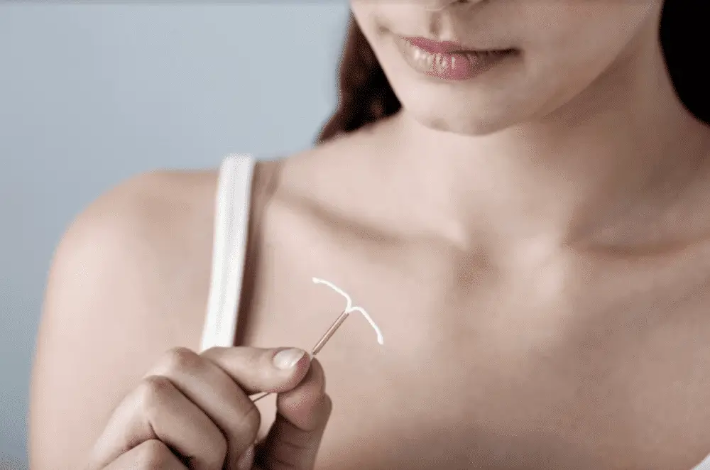 A woman in a white tank top holds an intrauterine device (IUD) contraceptive in her hand, close to her chest, with only the lower half of her face and torso visible.