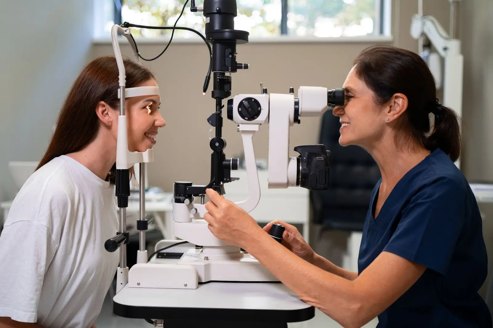 An eye doctor in scrubs examines a smiling young woman’s eyes using a slit lamp in a modern clinic. Both are facing each other, and the room is brightly lit with medical equipment in the background.