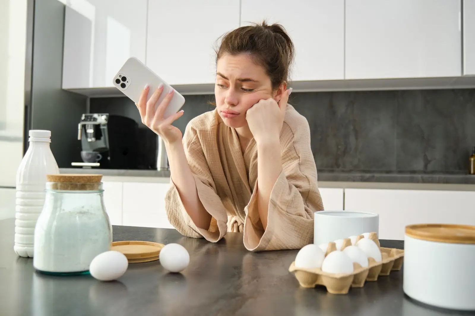 A woman in a robe leans on a kitchen counter, looking bored or frustrated at her phone. Eggs, a bottle of milk, flour, and other baking ingredients are on the counter in front of her. 