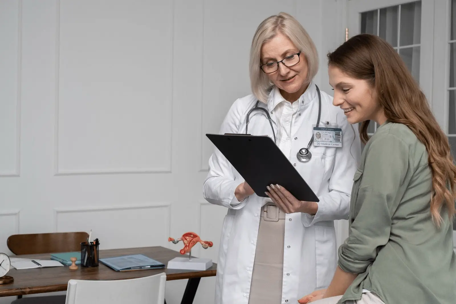 A female doctor with glasses discusses contraception treatments like Nexplanon to a smiling young woman sitting on an exam table in a medical office. A stethoscope hangs around the doctor’s neck and a laptop sits on the desk in the background.