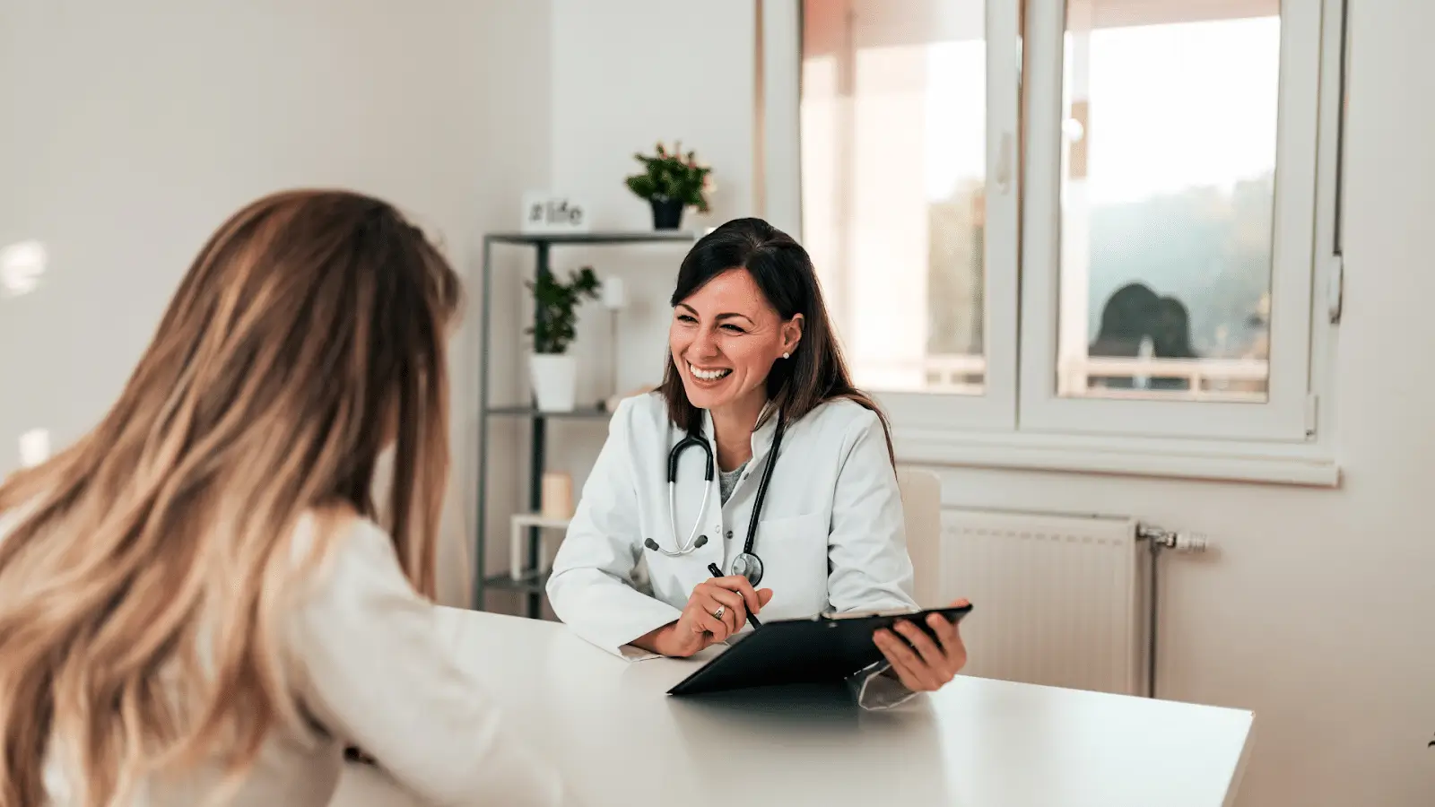 A smiling female doctor with a stethoscope talks to a patient across a desk in a bright, modern office, holding a tablet and taking notes.