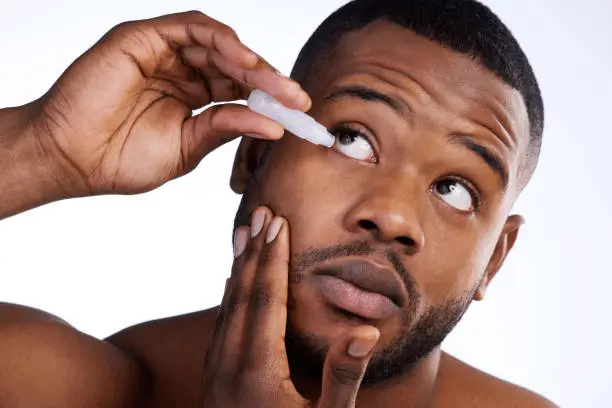 A man holds his lower eyelid down and applies eye drops to his right eye, looking upward with a focused expression. 