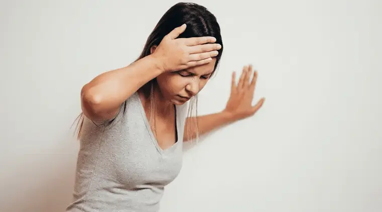 A woman with long hair holds her head with one hand and steadies herself against a wall with the other, appearing dizzy or unwell.