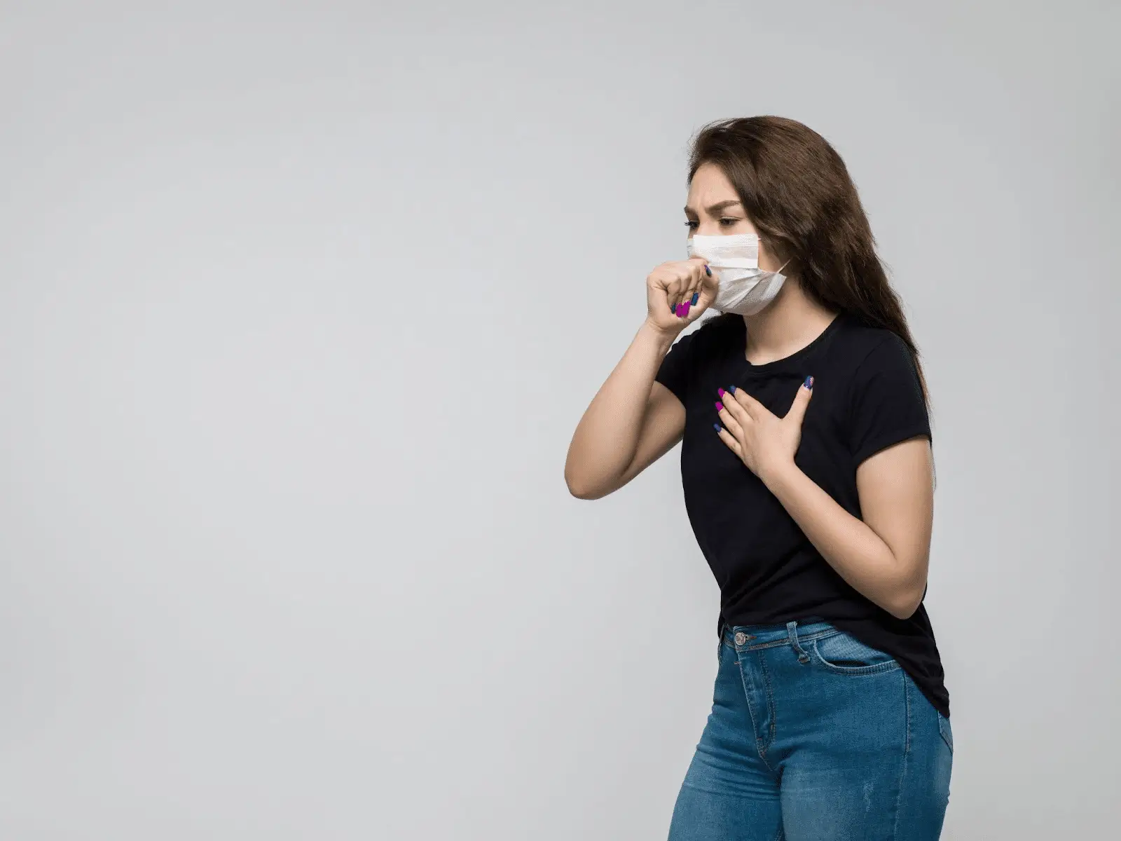 A young woman wearing a face mask coughs into her fist while holding her chest with her other hand. She is dressed in a black t-shirt and blue jeans, standing against a plain light gray background.