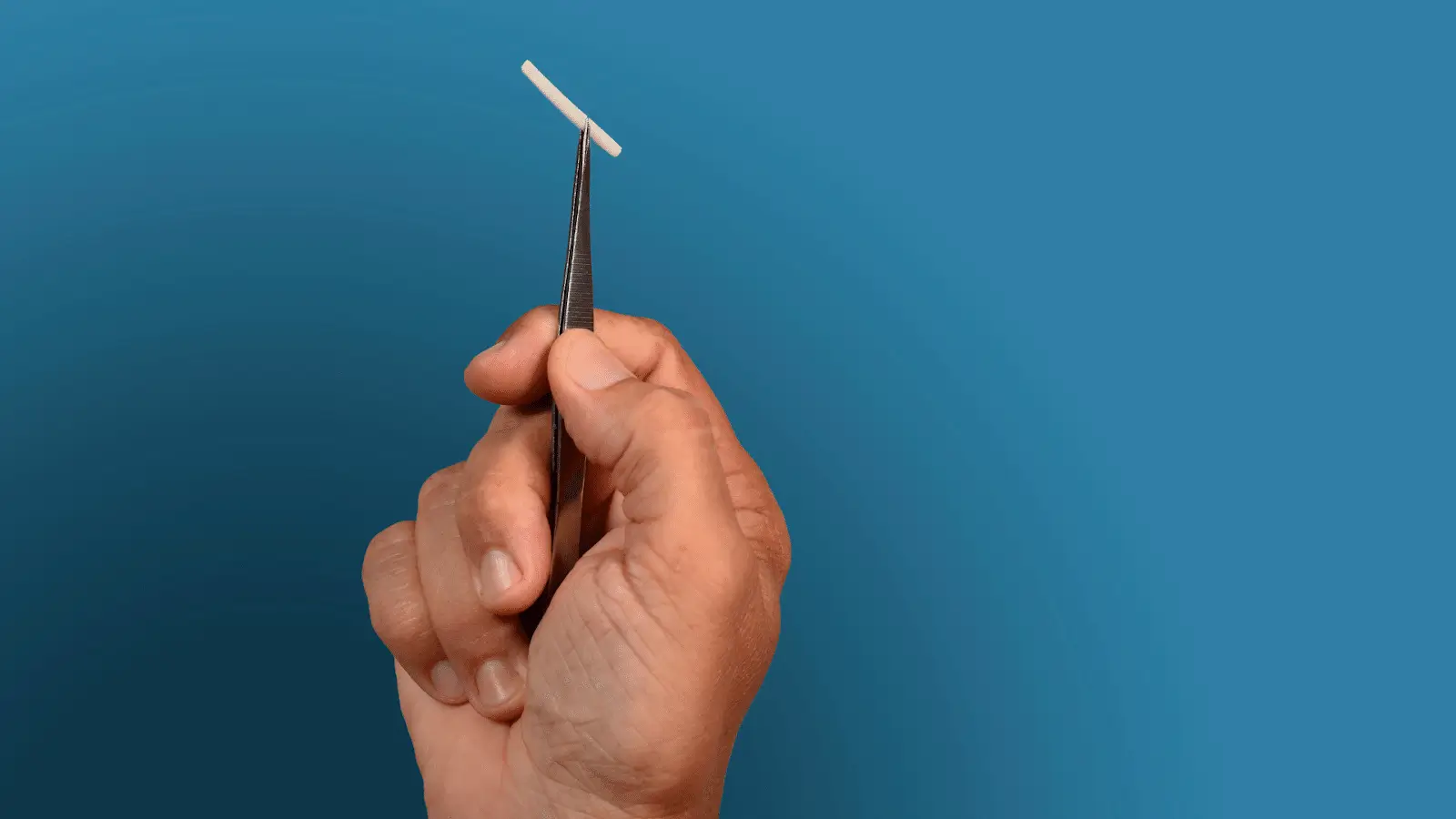 A hand holds forceps gripping a small, white, T-shaped intrauterine device (IUD) against a plain blue background.