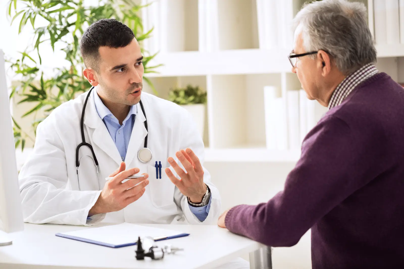 A doctor in a white coat and stethoscope sits at a desk, attentively talking to an older man in glasses and a purple sweater during a medical consultation.